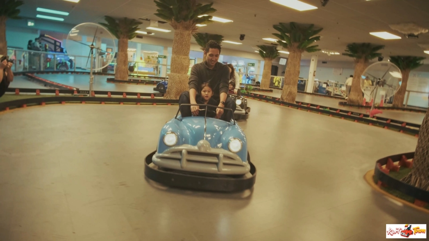 Two people ride in a blue bumper car on a track surrounded by artificial palm trees and large fans.