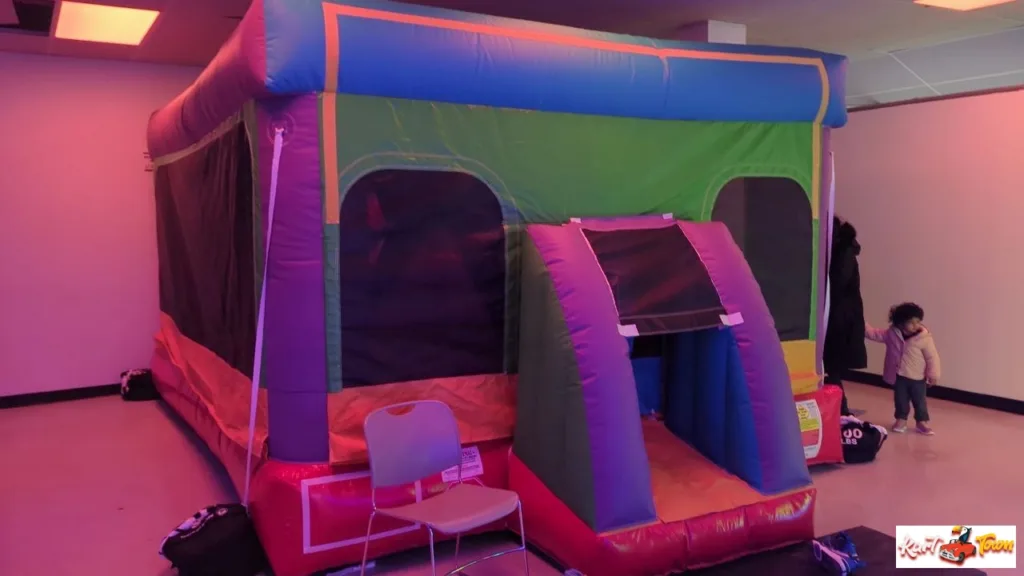 Colorful inflatable bounce house with mesh windows and a padded entrance ramp under pink lighting. A chair sits in front, and two people stand to the right.