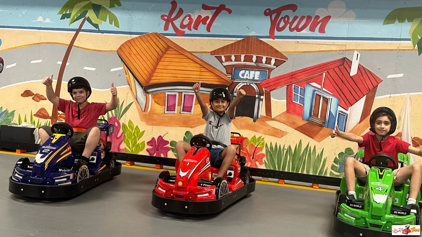 Three helmeted people give thumbs up in blue, red and green go-karts in front of a colorful mural reading “Kart Town.”