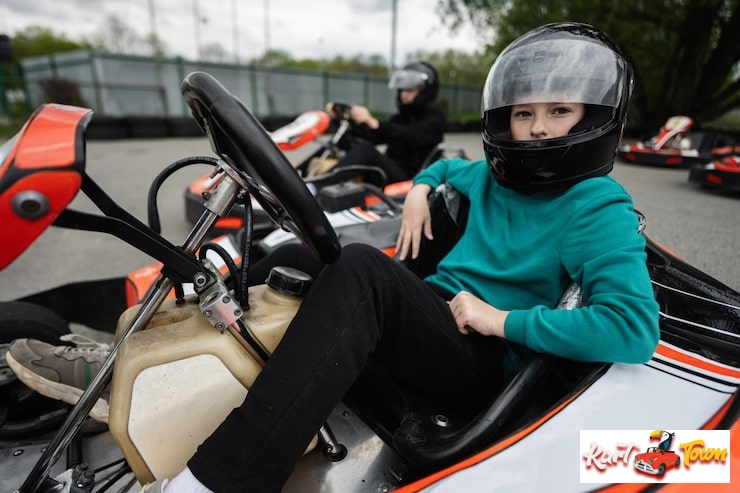 A child in a black helmet and teal sweatshirt sits casually in a white and black go-kart on an outdoor track, looking directly at the camera. Go Karting Weight Limit