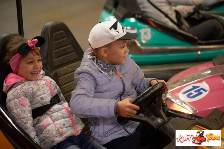 A boy with a cap is driving a bumper car while a younger girl with a headband laughs happily beside him on a ride.