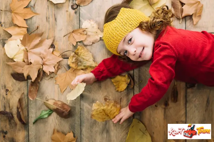 A happy young child wearing a yellow knit headband lying on a wooden floor with autumn leaves.