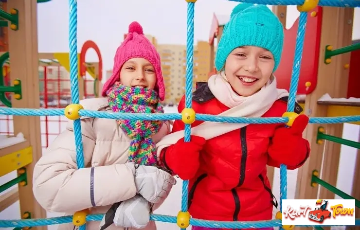 Two smiling girls in warm winter clothes peering through a colorful rope climbing structure.
