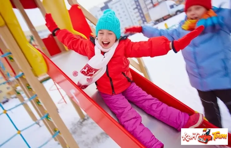 A cheerful girl in a red jacket and hat sliding down a red playground slide in the snow.