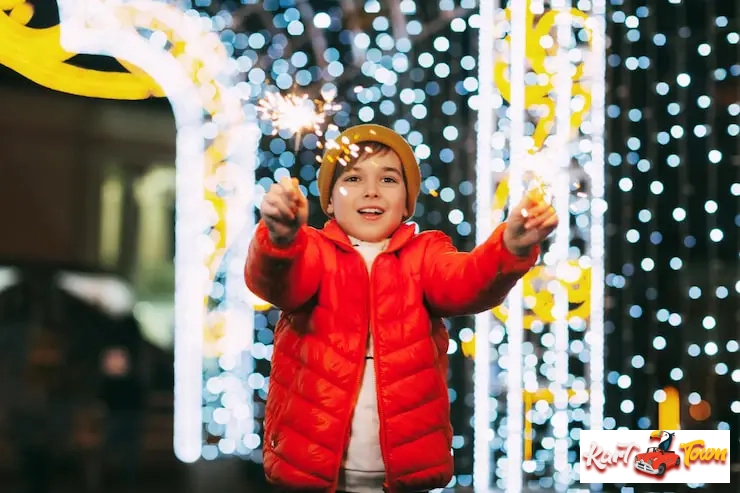 A boy wearing an orange puffer jacket and a yellow beanie smiles while holding sparklers.