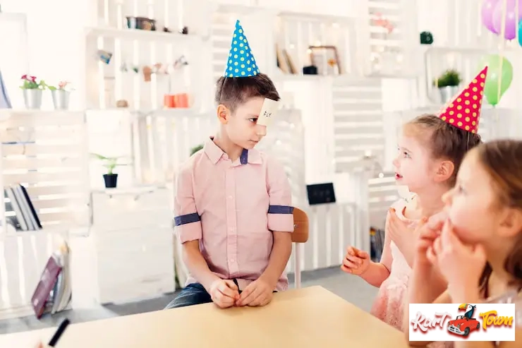 A boy wearing a party hat and a name-guessing game card on his forehead plays with two girls.
