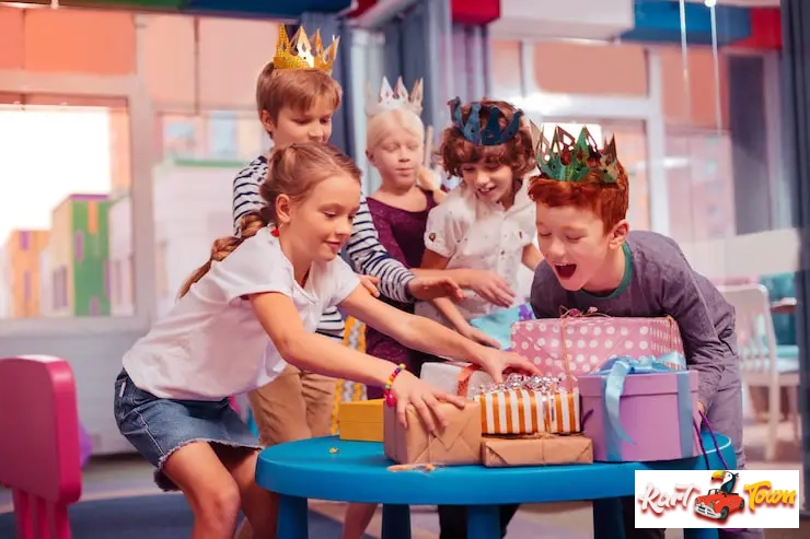 A group of children wearing birthday crowns excitedly reach for a stack of brightly gifts.