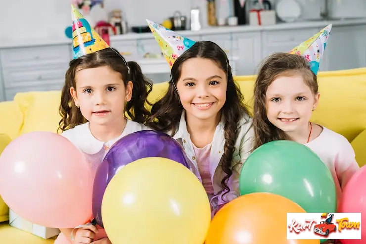 Three smiling girls wearing colorful party hats pose on a yellow sofa.