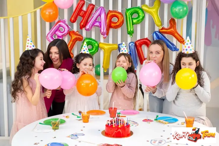 Adults and kids wearing party hats happily blowing up balloons around a birthday cake.