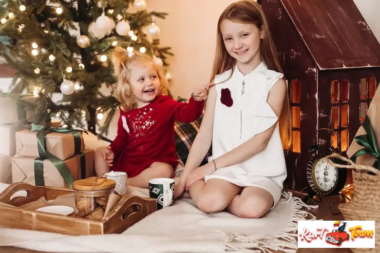 Two girls sitting on the floor by a Christmas tree and a toy house.