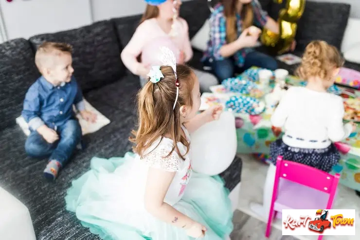 A young girl in a white dress sits on an indoor birthday celebration.