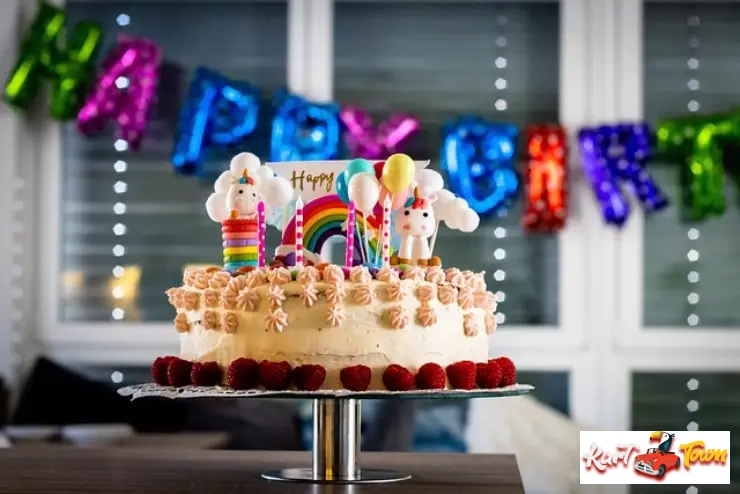 Unicorn and rainbow-themed birthday cake on a stand with "Happy Birthday" balloons behind it.