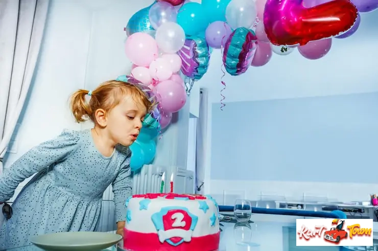 Young child blows out candles birthday cake under colorful balloons.