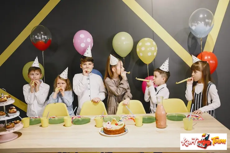 Six children standing in a row at a party table, wearing hats and blowing paper whistles.