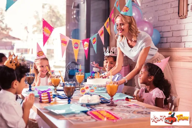 A woman serving a birthday cake to happy children at a decorated indoor party table.
