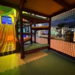 Yellow double slide beside padded climbing platforms, enclosed by black netting above interlocking foam floor mats.
