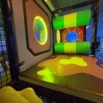 Netted play structure with green and yellow padded rollers, glowing oval windows, and a padded floor with colorful shapes.
