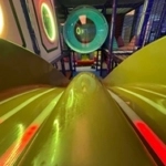 Yellow slide lanes beneath green-lit circular openings, with black netting and a colorful logo in the lower right.