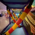 Net-enclosed play tunnel with crisscrossing red, yellow and teal padded poles, blue flooring and purple supports.