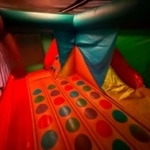 Colorful inflatable play structure with orange, green-spotted obstacles, red and teal walls, and a logo in the lower right.