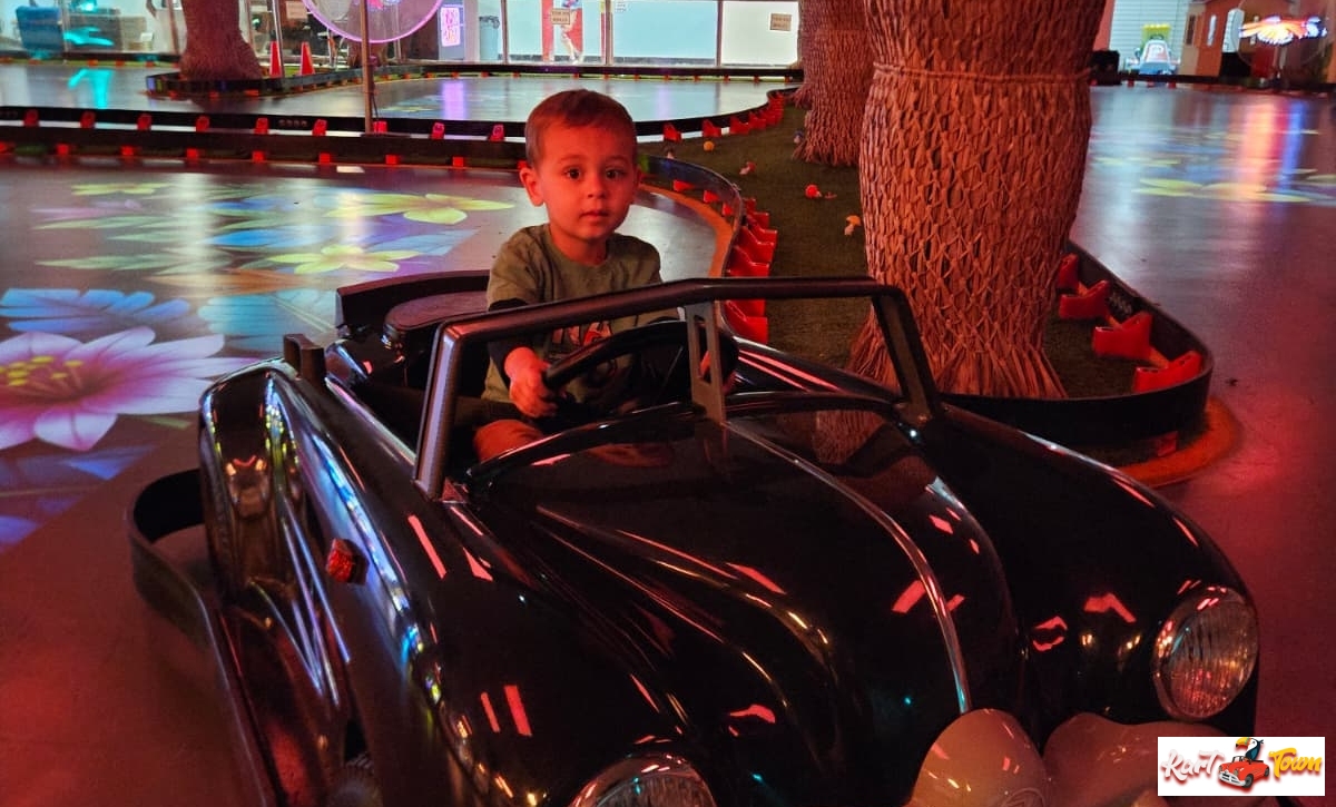 A child holds the steering wheel of a black classic ride-on car beside artificial palm trees and colourful floor projections.