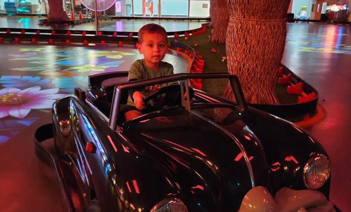 A child holds the steering wheel of a black classic ride-on car beside artificial palm trees and colourful floor projections.