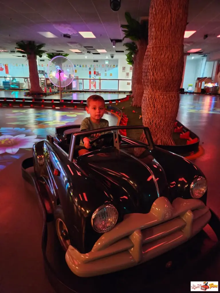 A child holds the steering wheel of a black classic ride-on car beside artificial palm trees and colourful floor projections.