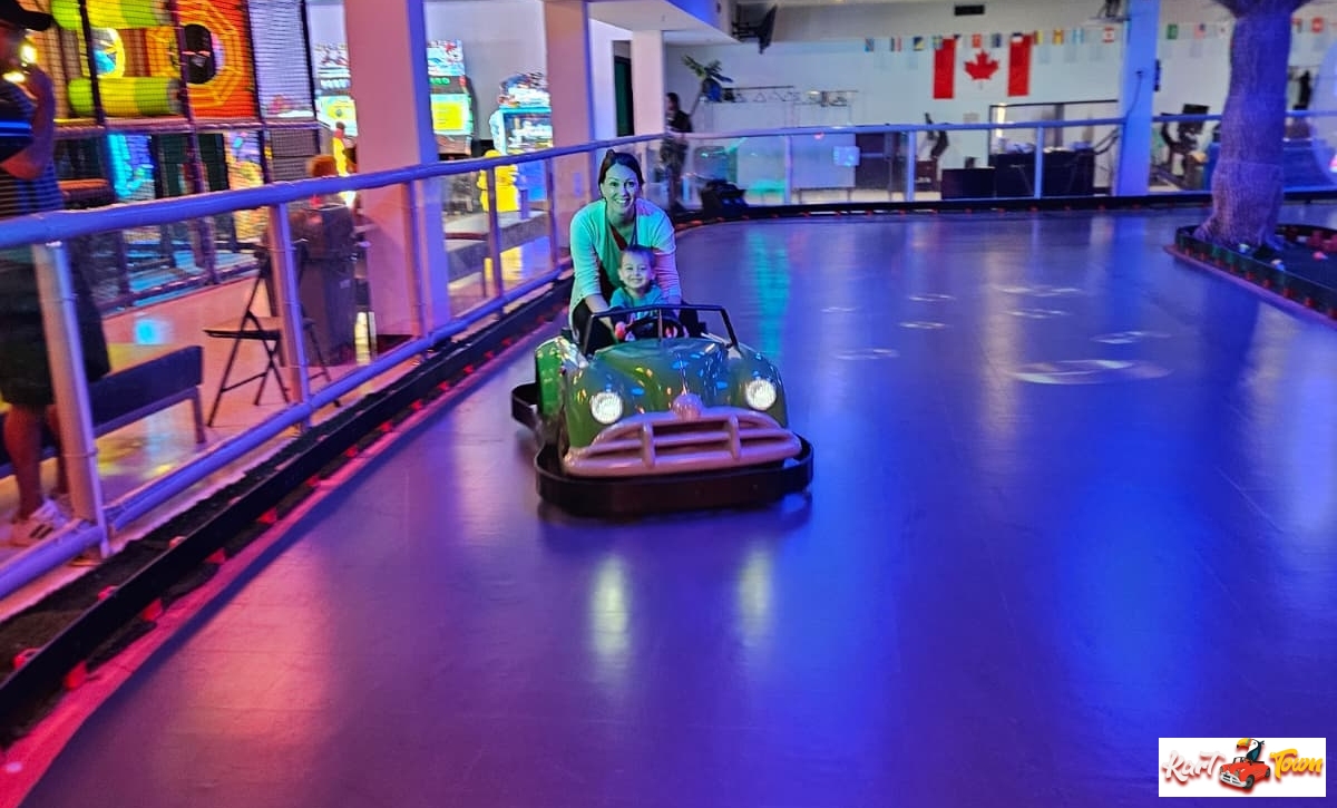 An adult and child sit in a green ride-on car on an indoor track under blue and purple lighting.