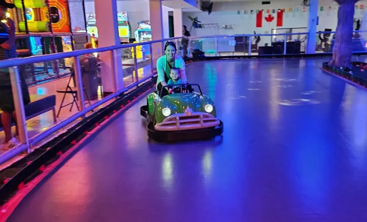 An adult and child sit in a green ride-on car on an indoor track under blue and purple lighting.