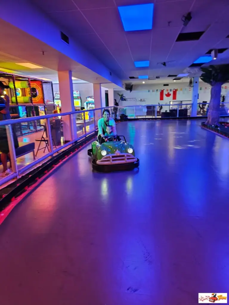 An adult and child sit in a green ride-on car on an indoor track under blue and purple lighting.