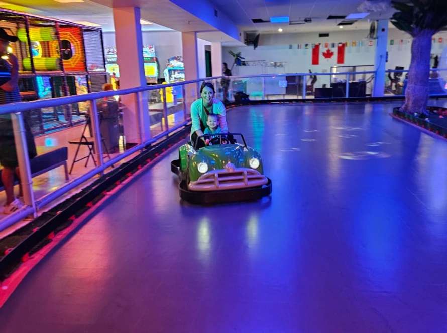 An adult and child sit in a green ride-on car on an indoor track under blue and purple lighting.