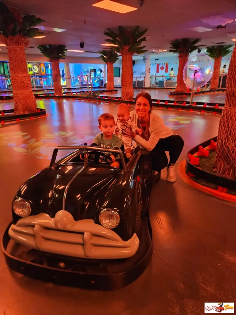 A child sits at the wheel of a black ride-on car while an adult holding a baby crouches beside it.