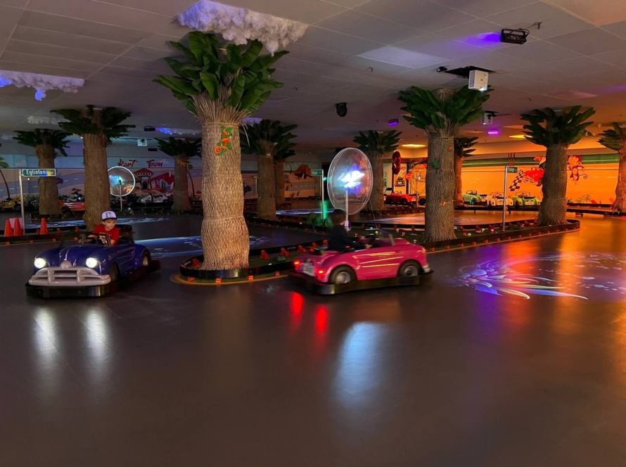 Two children driving purple and pink classic cars around the palm-tree indoor track