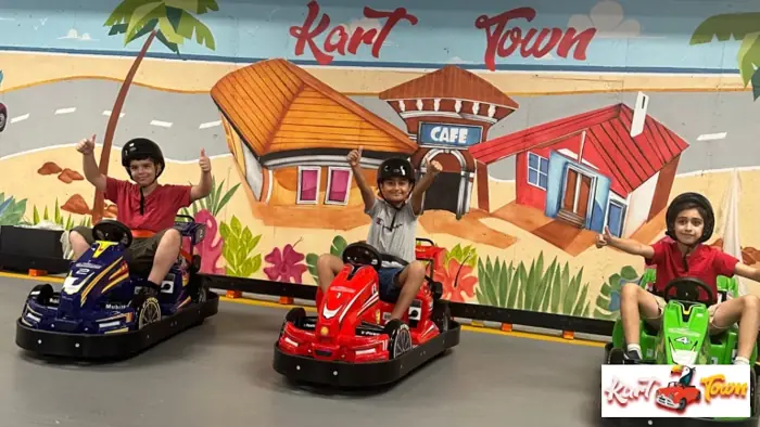 Three helmeted people give thumbs up in blue, red and green go-karts in front of a colorful mural reading “Kart Town.”