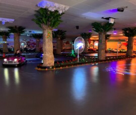 A driver rounds the illuminated palm-tree track in one of our shiny pink classic cars. Pink classic car driving around the palm-tree indoor track under sunset lighting