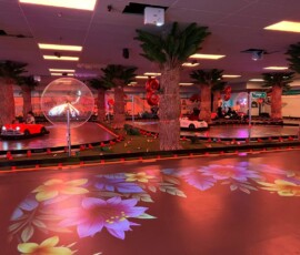 Neon lights, floral floor projections and karts in motion — the full Kart Town atmosphere on race day. Kart Town indoor track lit in neon red with karts racing, palm trees and floral projections
