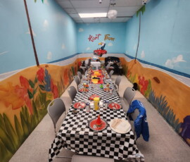 Celebrate in our Bird Room — a cheerful blue-sky mural with a fully set party table ready to go. Bird Room birthday party space with blue sky, flowers and a toucan driving a car mural at Kart Town Aurora